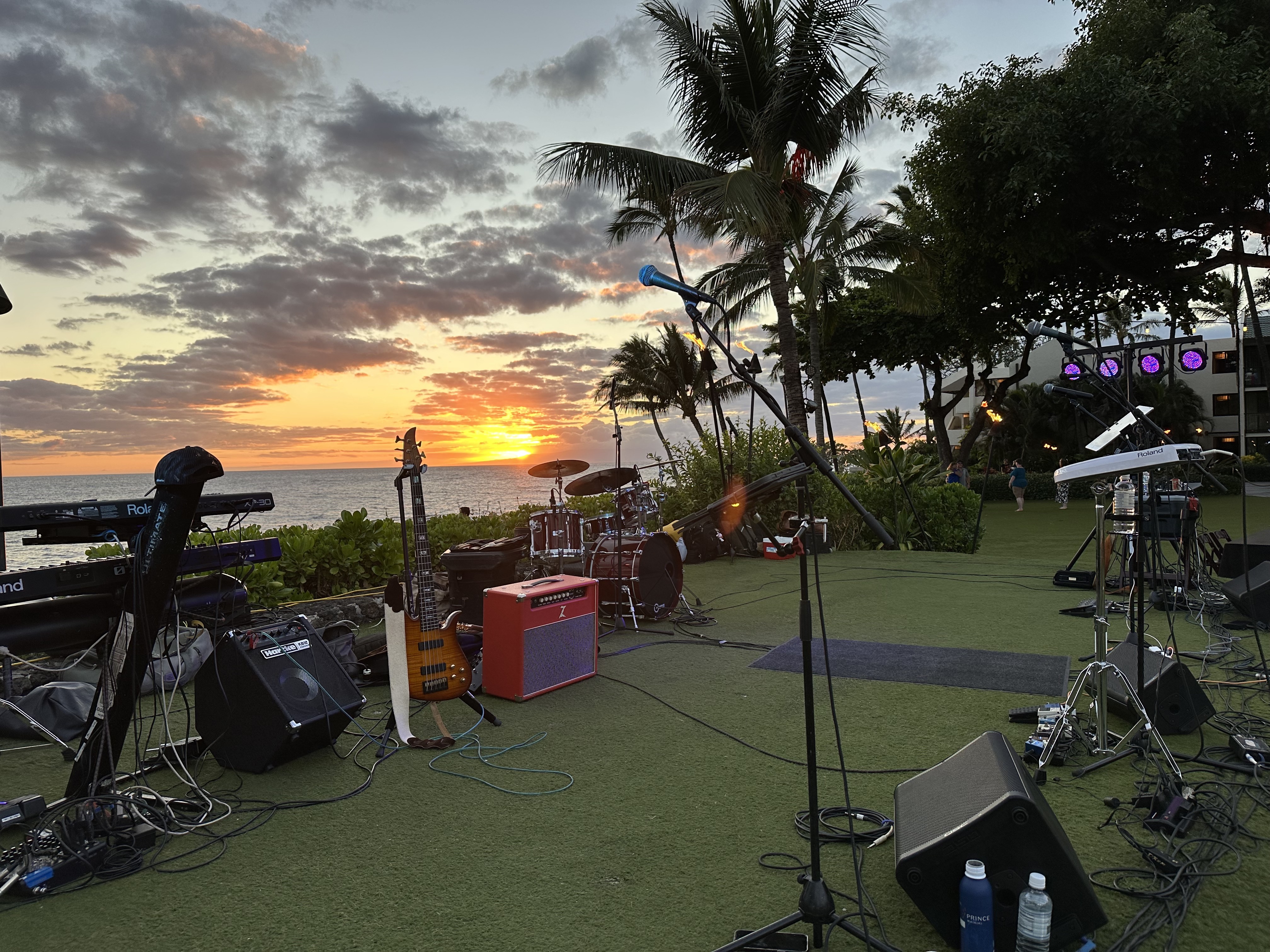 Backline speakers and stage gear silhouetted against a warm orange sunset at an outdoor event site